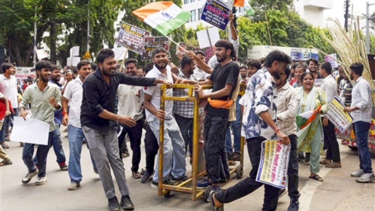 Large group of students and protesters marching on a road in Patna. Several protesters are carrying flags and placards as they participate in the demonstration against the state government. The march includes students walking alongside a wooden barricade-like structure, with buildings, trees and other people visible in the background.